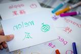 a thumb of a person holding an index card with green writing on the card with crayons in the background. 